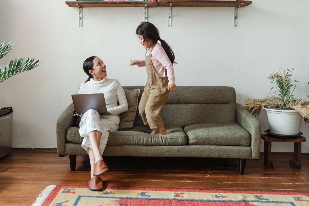A mum working on a laptop at home on a sofa while a young girl is clambouring on it